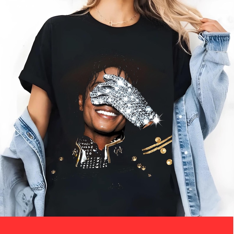 May include: Black t-shirt with a portrait of a smiling person, one eye covered by a sparkling, bejeweled glove. The person is wearing a black jacket with gold detailing. The image is a close-up, focusing on the face and upper body.