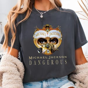 May include: Dark grey t-shirt with a gold and white graphic of Michael Jackson's eyes, the words "MICHAEL JACKSON" above and "DANGEROUS" below. The design is ornate and detailed.