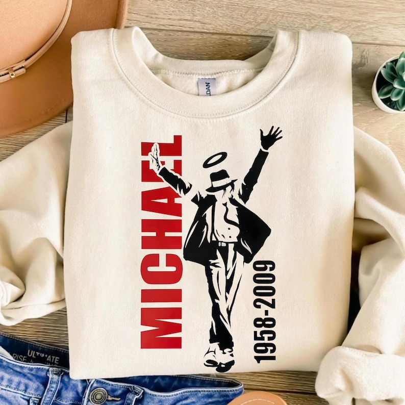 May include: A cream-coloured sweatshirt with a black and red graphic of a dancing figure. The name "MICHAEL" is printed vertically in red, with the dates "1958-2009" in black. The sweatshirt has a crew neck.