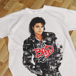 Pode incluir: Camiseta branca com um retrato de Michael Jackson. O design inclui a palavra "BAD" em vermelho, uma coroa e o n&uacute;mero "25". A camiseta tem mangas curtas e gola redonda.