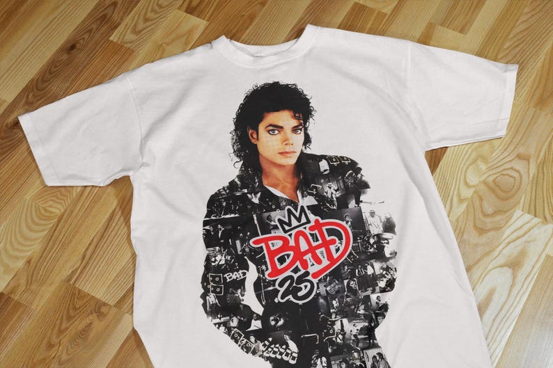 Pode incluir: Camiseta branca com um retrato de Michael Jackson. O design inclui a palavra "BAD" em vermelho, uma coroa e o n&uacute;mero "25". A camiseta tem mangas curtas e gola redonda.