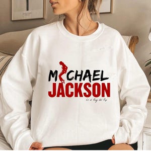 May include: White sweatshirt with the text "MICHAEL JACKSON" in black and red, and a red dancer silhouette. Below the name, the text "Es el Rey del Pop" is printed in black. The sweatshirt is a casual style.