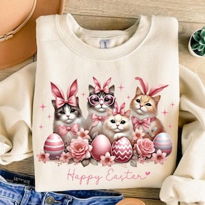 May include: Cream-colored sweatshirt featuring a graphic of four cats wearing pink bunny ears and bows, surrounded by Easter eggs and flowers. The words "Happy Easter" are printed in pink. The design is centered on the front of the shirt.