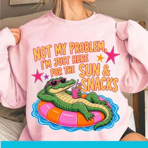 May include: A light pink sweatshirt with a graphic of a green alligator wearing sunglasses, floating on a pink and orange inner tube, holding a drink. Text reads "NOT MY PROBLEM, I'M JUST HERE FOR THE SUN & SNACKS."