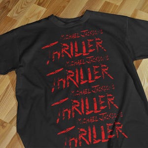 Pode incluir: Camiseta preta com a frase "MICHAEL JACKSON'S THRILLER" em vermelho, repetida v&aacute;rias vezes. O texto tem uma apar&ecirc;ncia desgastada, ligeiramente sangrenta. A camiseta est&aacute; sobre uma superf&iacute;cie de madeira.