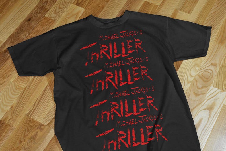 Pode incluir: Camiseta preta com a frase "MICHAEL JACKSON'S THRILLER" em vermelho, repetida v&aacute;rias vezes. O texto tem uma apar&ecirc;ncia desgastada, ligeiramente sangrenta. A camiseta est&aacute; sobre uma superf&iacute;cie de madeira.