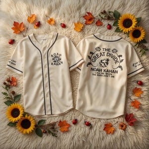 Può includere: Due maglie da baseball color crema con finiture e testo neri. Una maglia recita "NOAH KAHAN" e l'altra "THE GREAT DIVIDE NOAH KAHAN CALL ME WHEN THE BUGS DON'T DIE". Le maglie sono circondate da girasoli, foglie e coccinelle.