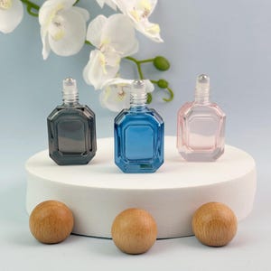 May include: Three small glass roller bottles with metallic roller balls, featuring an octagonal shape in black, blue, and light pink. The bottles are arranged in a row, each topped with a clear cap, showcasing a sleek and functional design for liquids.