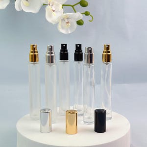 Puede incluir: Seis frascos de perfume cilíndricos de vidrio dispuestos en fila. Tres frascos son de vidrio esmerilado y tres de vidrio transparente. Cada uno cuenta con un atomizador en dorado, plateado o negro, con tapas a juego colocadas delante.