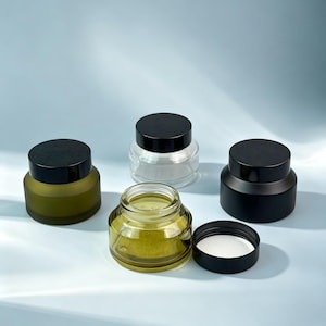 Op de afbeelding: Een verzameling van vier ronde cosmetische potjes met zwarte deksels. Eén potje is van helder glas, één is mat olijfgroen, één is massief matzwart en één is van transparant olijfgroen glas, getoond met het deksel verwijderd en een witte voering.