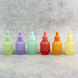 May include: Six colorful dropper bottles in a row. The bottles are pastel shades of green, purple, and blue, with red, orange, and yellow. Each bottle has a matching colored dropper top. The bottles are on a gray surface.