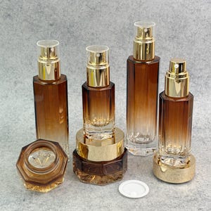 May include: An assortment of brown and gold cosmetic bottles and jars. The bottles have gold spray tops and clear caps. The jars have gold lids. The glass has a gradient effect, transitioning from brown to clear.