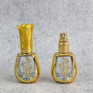 Op de afbeelding: Twee decoratieve parfumflesjes met goudkleurige accenten. De flesjes hebben een heldere glazen body met een gouden bloemenontwerp. Eén flesje heeft een bloemvormige dop, de andere een verstuiver.