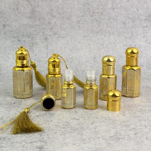 May include: A collection of gold-colored perfume bottles with intricate etched designs. Some bottles have gold caps, while others have rollerball applicators. A gold tassel is attached to some of the bottles. The bottles are arranged on a gray surface.