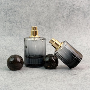 May include: Two glass perfume bottles with gold-colored spray tops and dark gray ombre coloring. Two dark brown, spherical bottle stoppers are also present. The bottles have a ribbed design and are displayed on a gray surface.