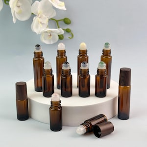 May include: A collection of amber glass roller bottles with dark wood-grain caps and various gemstone roller balls. The bottles are arranged on a white circular platform, with some standing upright and one lying horizontally in the foreground.
