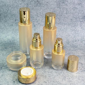 May include: A collection of cosmetic bottles and jars with gold-colored caps. The bottles are clear with a frosted, peach-colored gradient. The set includes various sizes of spray bottles and a small jar with a screw-on lid.