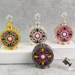 Puede incluir: Frascos de perfume ornamentados en tonos dorados, plateados y bronce, cada uno con un diseño decorativo con incrustaciones de joyas. Los frascos presentan una perla central y un tapón de cristal. Se exhiben sobre una superficie blanca.