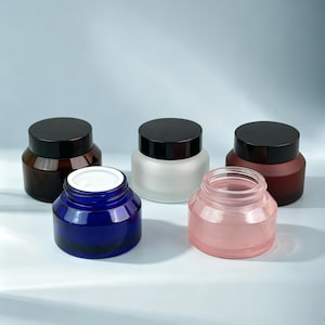 May include: Five cosmetic jars with black lids. Three jars are closed, featuring dark brown, frosted white, and deep red glass. Two jars are open, one made of translucent blue glass with a white inner seal and one made of clear pink glass.