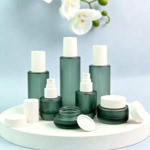 May include: A collection of frosted dark green glass cosmetic containers with matte white caps and pump dispensers. The set includes various sizes of cylindrical bottles and round jars, some with lids removed to show the pump and open jar tops.