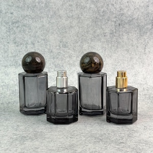 May include: Four gray glass perfume bottles with different shaped bases and tops. Two bottles have a dark brown, spherical cap. The other two have silver and gold spray tops. The bottles are on a gray surface.