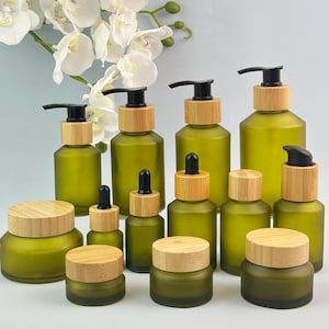 Puede incluir: Una colección de recipientes cosméticos de vidrio esmerilado verde oliva con tapas de madera clara y dispensadores de bomba o gotero negros. El conjunto incluye varios tamaños de frascos y botellas, dispuestos con flores de orquídea blancas.