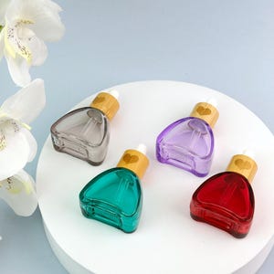 May include: Four triangular glass dropper bottles in gray, purple, teal, and red. Each bottle features a light-colored wooden cap with a small heart icon engraved on the top, paired with a white rubber dropper bulb for dispensing liquids.
