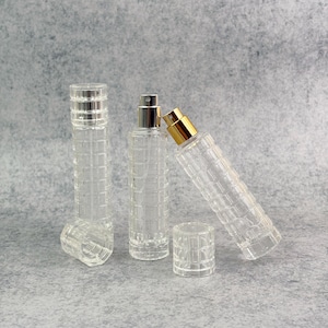 Puede incluir: Botellas de perfume de vidrio transparente con atomizadores plateados y dorados. Las botellas tienen un patrón texturizado en forma de rejilla. Una botella está sin tapón, otra está inclinada.