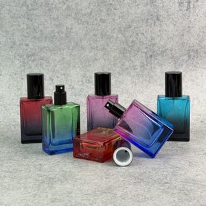 May include: A collection of rectangular glass perfume bottles with black spray nozzles and caps. The bottles display a gradient of colors, including red, green, blue, and purple. The bottles are arranged on a gray surface.