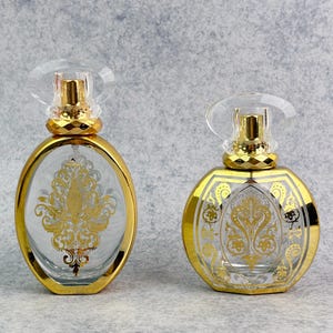 Puede incluir: Dos frascos de perfume ornamentados. Uno es ovalado, el otro redondo. Ambos tienen detalles dorados y vidrio transparente con diseños de filigrana dorada. Los frascos tienen tapones transparentes y redondeados y tapas doradas. El fondo es gris moteado.