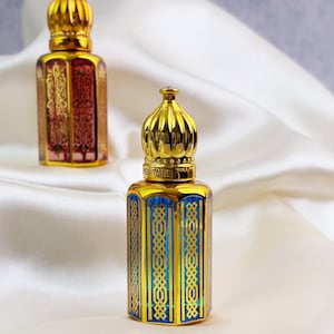 Op de afbeelding: Twee sierlijke parfumflesjes met goudkleurige doppen en bodems. Eén flesje heeft een rood-gouden ontwerp, de andere een blauw-gouden ontwerp. De flesjes staan op een witte, zijdeachtige ondergrond.