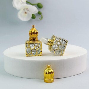 May include: Two small square glass perfume bottles with gold-colored metal honeycomb overlays and matching gold-colored dome-shaped caps. One bottle stands upright with its cap on, while the other lies on its side with the cap removed.