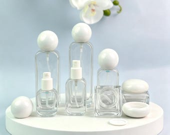 May include: A collection of clear glass cosmetic containers featuring white spherical caps and white pump sprayers. The set includes various sizes of rectangular bottles and small jars with matching white lids, arranged on a white circular base.