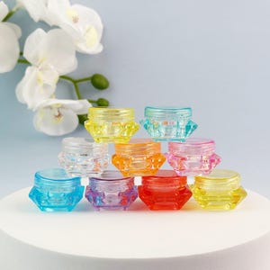 Peut inclure: Une collection de petits pots cosmétiques translucides en forme de diamant empilés en pyramide. Les contenants présentent des bords à facettes et sont disponibles dans une variété de couleurs vives, notamment jaune, bleu, orange, rose, violet et rouge.
