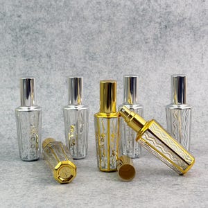 Pode incluir: Uma coleção de frascos de perfume de vidro com detalhes prateados e dourados. Os frascos têm formato retangular com padrões decorativos e borrifadores metálicos. Alguns frascos são prateados, outros dourados.