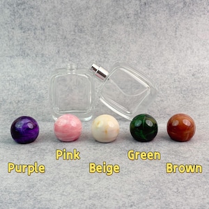 May include: Clear glass perfume bottles with silver sprayers, next to five decorative, spherical perfume bottle stoppers in purple, pink, beige, green, and brown. The colours are labelled below each stopper.