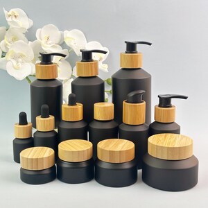 May include: A collection of matte black cosmetic containers with light wood grain lids and pump dispensers. The set includes various sizes of bottles with pumps, dropper bottles, and jars with screw-on lids, arranged in a group against a light background.
