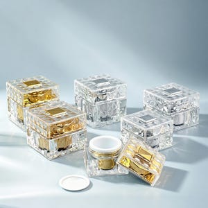 May include: Clear, faceted cosmetic jars with gold and silver accents. The jars are square with a diamond-like pattern and a small square on the lid. Some jars have gold-colored interiors, while others are silver. A white lid and a gold-rimmed container are also present.
