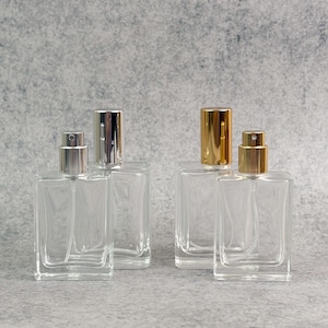 Puede incluir: Cuatro frascos de perfume de vidrio transparente con pulverizadores plateados y dorados. Los frascos son rectangulares y vacíos, con una boquilla pulverizadora. El fondo es gris claro.