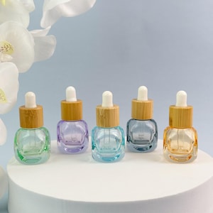 May include: Five small, square-shaped glass dropper bottles arranged in a row. Each bottle features a light-colored bamboo collar and a white rubber dropper bulb. The glass bases are tinted in distinct colors: light green, purple, blue, grey, and amber.