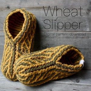 Op de afbeelding: Een paar handgemaakte tarwekleurige pantoffels met olijfgroene accenten. De pantoffels zijn gemaakt van dik garen en hebben een textuur, geweven ontwerp. De tekst "Wheat Slipper" en "Digital pattern download" zijn zichtbaar.