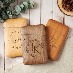 Könnte beinhalten: Drei rechteckige Holzetuis mit Klappdeckeln. Das linke Etui ist aus hellem Holz mit dem Text "HELEN JAMES" und einem Blumenmuster. Das mittlere Etui ist aus dunklem Holz mit den Initialen "JK" in einem Blumenkranz. Das rechte Etui ist aus mittelbraunem Holz mit dem Buchstaben "J" und "Erica".