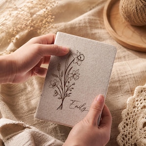 May include: A rectangular notebook with a light beige, textured fabric cover. The front features a dark brown line art illustration of a bouquet of daffodils and wildflowers, with the name Emily written in a cursive script below the flowers.