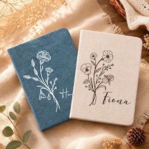 May include: Two rectangular notebooks with fabric covers. The left notebook is denim blue with a white line-art floral illustration. The right notebook is beige with a dark brown line-art floral illustration and the name Fiona printed in script.