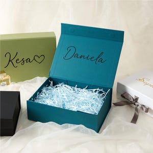 Puede incluir: Caja de regalo turquesa abierta con el nombre "Daniela" en cursiva. La caja está llena de papel triturado azul claro. Se ven otras cajas, incluida una caja verde con "Kesa" y un corazón, y una caja blanca con un lazo.