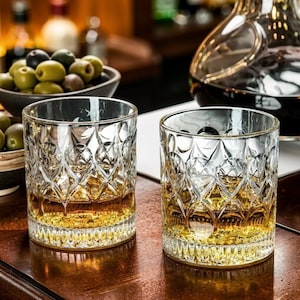 Puede incluir: Dos vasos de whisky de cristal con un diseño de diamantes, llenos de líquido ámbar. Los vasos están sobre una superficie de madera, con un cuenco de aceitunas y una jarra en el fondo. Los vasos miden unos 9 cm de alto.