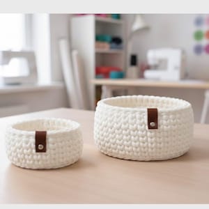 May include: Two white crochet baskets of different sizes sit on a light-colored wooden surface. Each basket has a brown leather strap with a silver button. The baskets are round and have a textured appearance.
