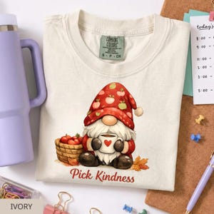 May include: A cream-colored t-shirt featuring a graphic of a gnome wearing a red hat with apple patterns, holding a white mug with a red heart. Next to the gnome is a basket of red apples. The text "Pick Kindness" is printed below the gnome.