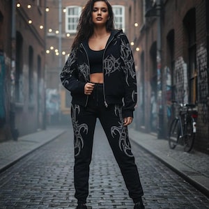 Op de afbeelding: Zwarte hoodie met rits en bijpassende joggingbroek met een tribaal ontwerp in een lichtere kleur. De outfit wordt gedragen met een zwarte crop top en laarzen. De achtergrond is een steeg met bakstenen en lichtslingers.