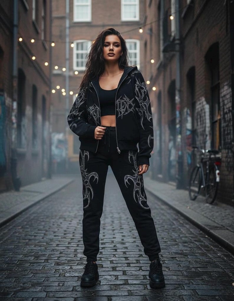 Op de afbeelding: Zwarte hoodie met rits en bijpassende joggingbroek met een tribaal ontwerp in een lichtere kleur. De outfit wordt gedragen met een zwarte crop top en laarzen. De achtergrond is een steeg met bakstenen en lichtslingers.